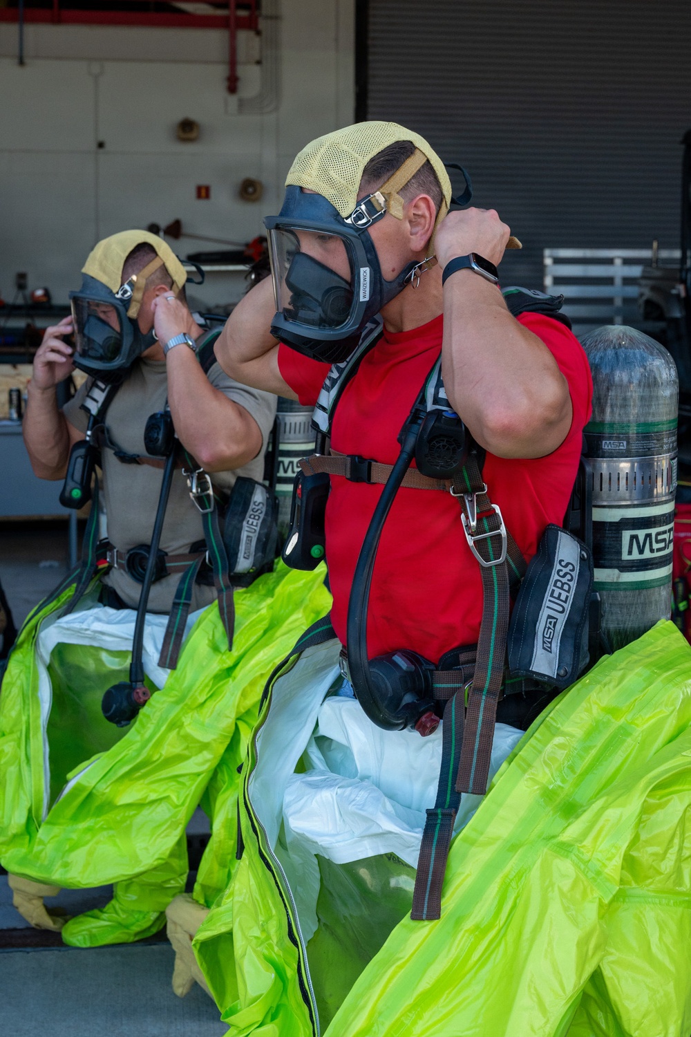Two individuals are donning MSA SCBA (Self-Contained Breathing Apparatus) equipment, including full-face respirator masks and air cylinders, in what appears to be a fire station or emergency response facility. Both are wearing yellow Nomex hoods and are in the process of adjusting their mask straps. Bright yellow-green hazmat or protective suits are visible pooled around their lower bodies, suggesting they are suiting up for a hazardous materials or firefighting exercise.