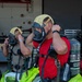 Two individuals are donning MSA SCBA (Self-Contained Breathing Apparatus) equipment, including full-face respirator masks and air cylinders, in what appears to be a fire station or emergency response facility. Both are wearing yellow Nomex hoods and are in the process of adjusting their mask straps. Bright yellow-green hazmat or protective suits are visible pooled around their lower bodies, suggesting they are suiting up for a hazardous materials or firefighting exercise.