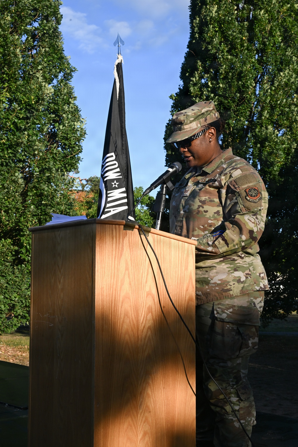 A U.S. Air Force service member in OCP (Operational Camouflage Pattern) uniform is speaking at a podium during an outdoor ceremony. A POW/MIA flag is prominently displayed beside the wooden lectern. The setting appears to be a formal military memorial or commemorative event held outdoors.