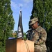 A U.S. Air Force service member in OCP (Operational Camouflage Pattern) uniform is speaking at a podium during an outdoor ceremony. A POW/MIA flag is prominently displayed beside the wooden lectern. The setting appears to be a formal military memorial or commemorative event held outdoors.
