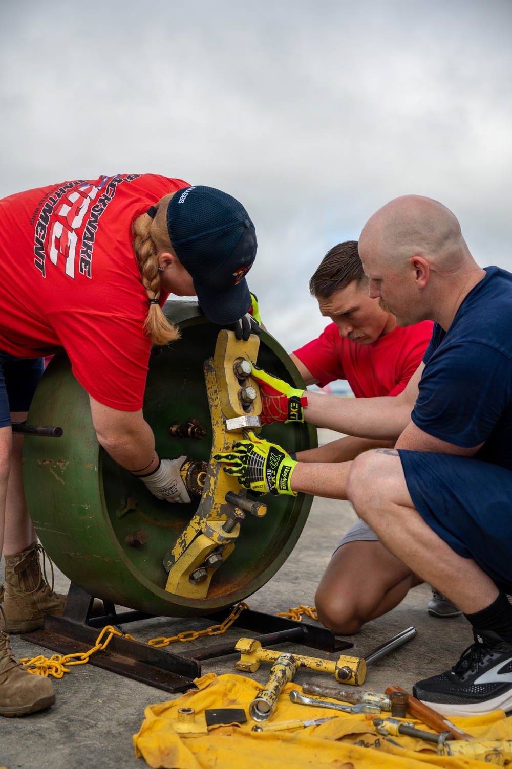 Three individuals are collaboratively working on a large green cylindrical object with yellow mechanical components, likely a pipe flange or valve assembly. They are using various hand tools and wearing protective gloves while performing what appears to be a maintenance or rescue training exercise. Tools including wrenches, bolts, and chains are laid out on a yellow cloth on the ground nearby.