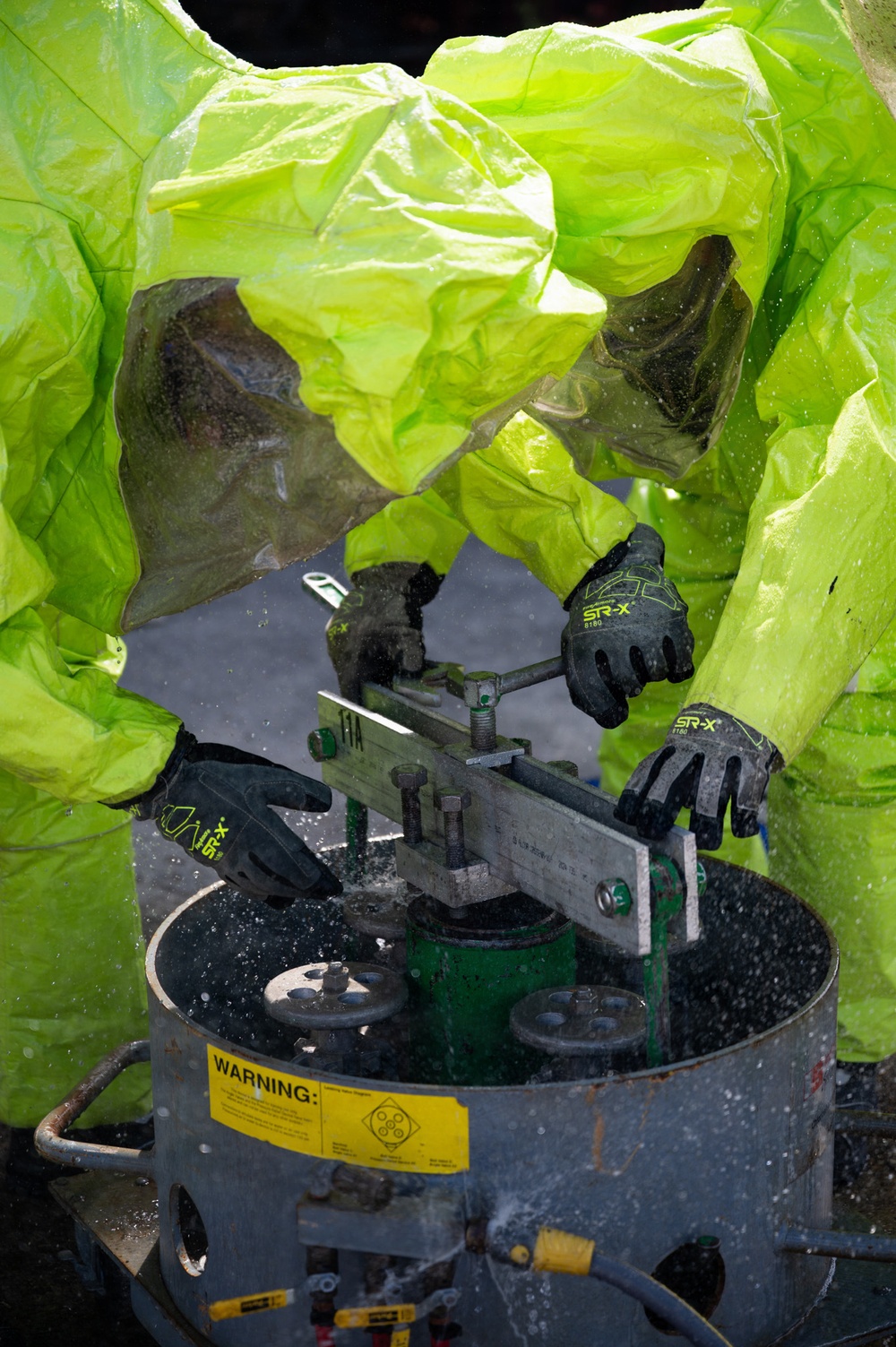 Two individuals wearing bright lime-green hazmat/chemical protective suits are working on industrial equipment. They are manipulating what appears to be a valve or pipe fitting assembly on a cylindrical metal container marked with a warning label. The scene suggests a hazardous materials response or industrial maintenance operation requiring full chemical protection.
