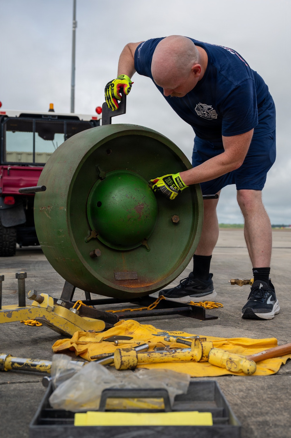 A bald man wearing a navy blue t-shirt, shorts, and high-visibility yellow impact-resistant gloves is leaning over a large green metal wheel or disc mounted on a stand on an airfield tarmac. He appears to be inspecting or manipulating the central hub assembly of the object. Various yellow-colored tools, chains, and equipment are spread on the ground nearby.