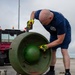 A bald man wearing a navy blue t-shirt, shorts, and high-visibility yellow impact-resistant gloves is leaning over a large green metal wheel or disc mounted on a stand on an airfield tarmac. He appears to be inspecting or manipulating the central hub assembly of the object. Various yellow-colored tools, chains, and equipment are spread on the ground nearby.