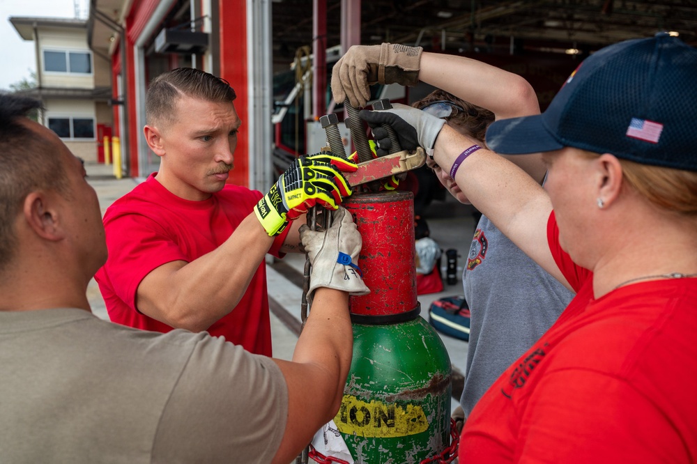 A group of firefighters or emergency responders are working together to manipulate the valve or regulator on a large green compressed gas cylinder outside a fire station. The central figure, wearing a red shirt and yellow impact-resistant gloves, is focused intently on the task. Multiple individuals are collaborating on the equipment, suggesting a training exercise or maintenance procedure.
