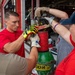 A group of firefighters or emergency responders are working together to manipulate the valve or regulator on a large green compressed gas cylinder outside a fire station. The central figure, wearing a red shirt and yellow impact-resistant gloves, is focused intently on the task. Multiple individuals are collaborating on the equipment, suggesting a training exercise or maintenance procedure.