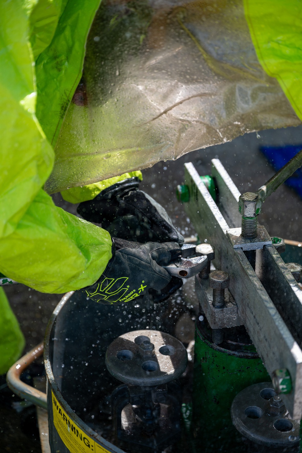 A worker wearing bright neon yellow/green protective gear and black gloves is performing mechanical work on industrial equipment. The gloves are branded 'FireArmor SRX' and the worker appears to be manipulating a bolt or fastener on a metal assembly. Water droplets are visible throughout the scene, suggesting a wet working environment.