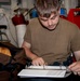 A young man wearing a olive/tan t-shirt is seated in what appears to be a military or industrial workspace, focused on opening or reviewing a white three-ring binder. The environment behind him includes cables, equipment, red containers, and a stool, consistent with a ship or facility workspace. A brown duffel bag is visible to his left.