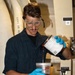 A young male worker wearing safety goggles and blue nitrile gloves is carefully pouring an amber/orange liquid from a white paint can into a smaller clear measuring container. He is dressed in a dark navy work uniform and appears to be in an industrial or naval vessel environment. The background shows large white pipes or ducts with stenciled text reading 'RAST DK DR'.