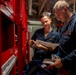Two naval personnel in dark blue coveralls are reviewing printed documentation inside what appears to be a ship's interior compartment. They are standing near a red equipment rack or panel, with one person holding papers and the other examining components. The setting is a confined, industrial shipboard space with visible piping, cables, and labeled equipment.