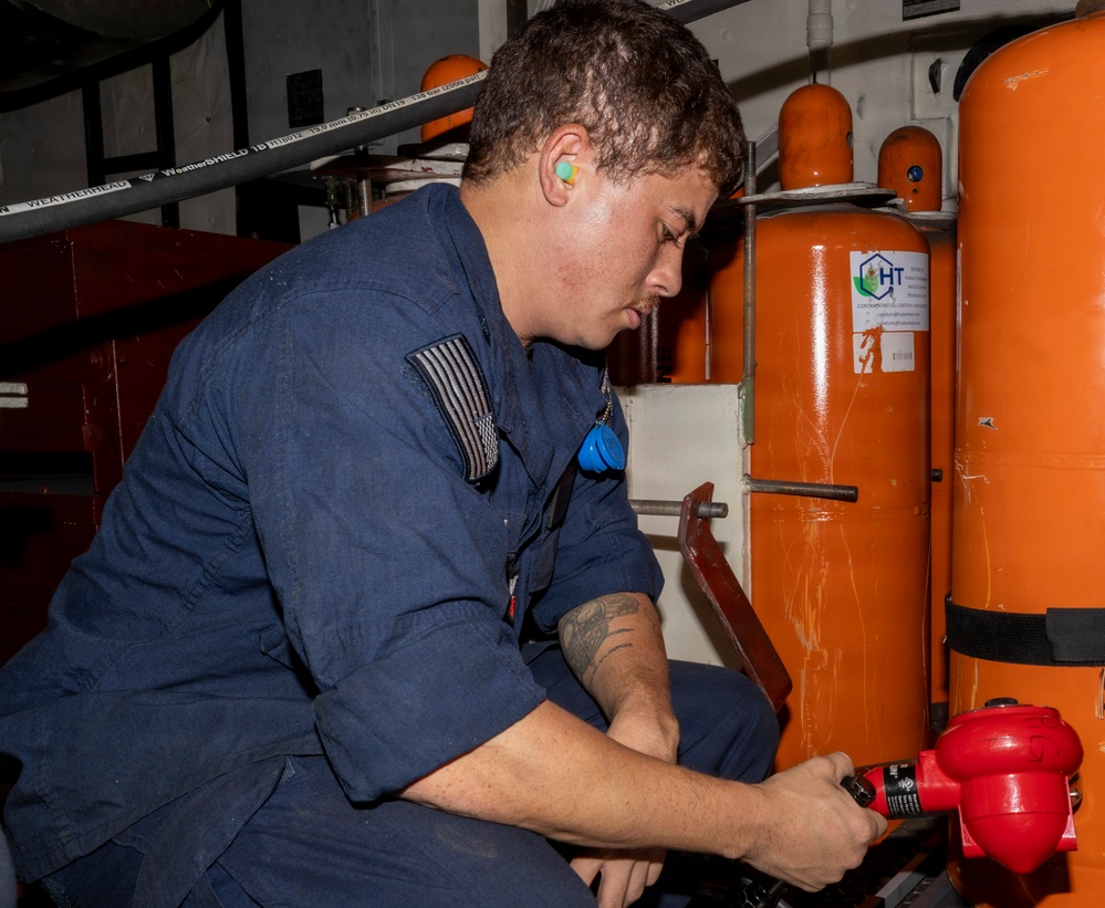A military or naval personnel member in blue coveralls is working on what appears to be fire suppression or pressurized gas equipment in an industrial or shipboard environment. The individual is crouching and manipulating a red valve or fitting connected to large orange pressurized cylinders. The setting appears to be a mechanical or utility space aboard a vessel or military installation.