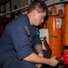 A military or naval personnel member in blue coveralls is working on what appears to be fire suppression or pressurized gas equipment in an industrial or shipboard environment. The individual is crouching and manipulating a red valve or fitting connected to large orange pressurized cylinders. The setting appears to be a mechanical or utility space aboard a vessel or military installation.