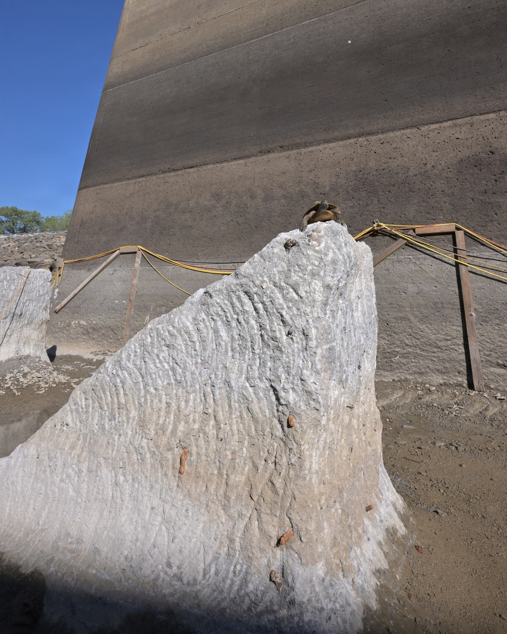 The image shows a large, rough-textured stone or concrete monolith in the foreground, positioned at the base of a massive smooth concrete wall or dam structure. Yellow safety ropes are strung between metal support frames in the background, suggesting an active construction or excavation site. A small bird is perched at the top of the rough stone formation.