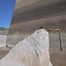 The image shows a large, rough-textured stone or concrete monolith in the foreground, positioned at the base of a massive smooth concrete wall or dam structure. Yellow safety ropes are strung between metal support frames in the background, suggesting an active construction or excavation site. A small bird is perched at the top of the rough stone formation.