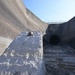 The image shows a large concrete dam or spillway structure photographed from a low angle, revealing a circular tunnel opening or outlet portal at its base. A rough-textured concrete or stone block is prominently visible in the foreground, with a small turtle resting on top of it. The structure appears to be in a dewatered or low-water condition, with wet ground, hoses, and construction equipment visible at the base.