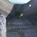 A turtle is perched on a white concrete or stone block in the foreground, while the background shows a large concrete structure — likely a dam spillway or drainage basin — that is currently drained and under maintenance or construction. A large circular tunnel opening is visible in the curved concrete wall, and construction hoses and equipment are scattered across the wet, muddy floor. The contrast between the small wildlife subject and the massive industrial infrastructure is a notable compositional element.