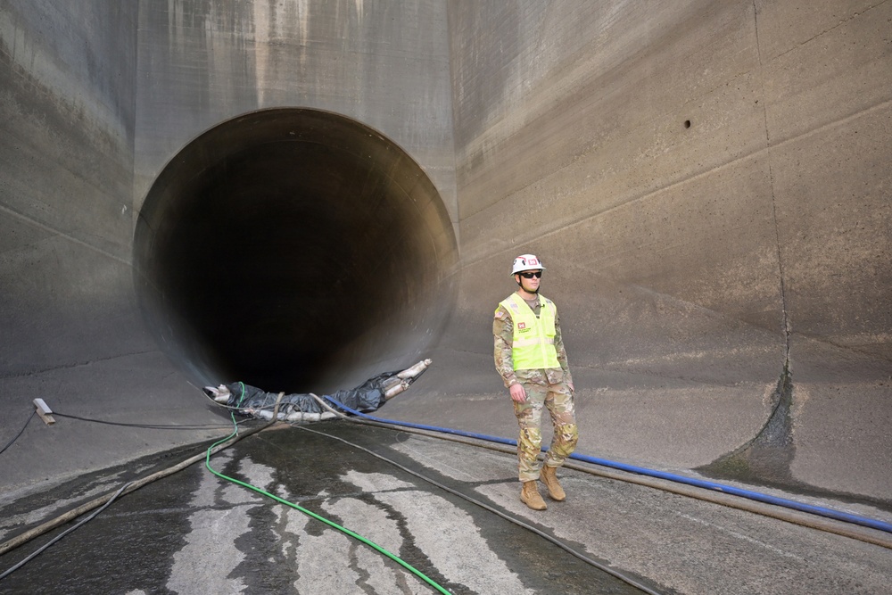 A military personnel member wearing camouflage uniform, high-visibility vest, white hard hat, and sunglasses stands in front of a large circular concrete tunnel or culvert opening. The setting appears to be a dam, spillway, or large water control infrastructure facility. Various hoses and cables are visible on the wet concrete floor, suggesting ongoing inspection or maintenance work.