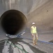 A military personnel member wearing camouflage uniform, high-visibility vest, white hard hat, and sunglasses stands in front of a large circular concrete tunnel or culvert opening. The setting appears to be a dam, spillway, or large water control infrastructure facility. Various hoses and cables are visible on the wet concrete floor, suggesting ongoing inspection or maintenance work.