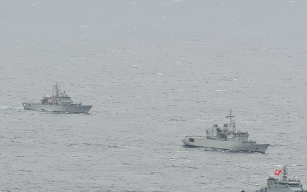 Three naval vessels are sailing in formation across open ocean under overcast, hazy conditions. The ships appear to be gray-painted military vessels of varying sizes, with the closest vessel bearing the hull number 'P813' and a red flag. The formation suggests a coordinated naval exercise or patrol operation.