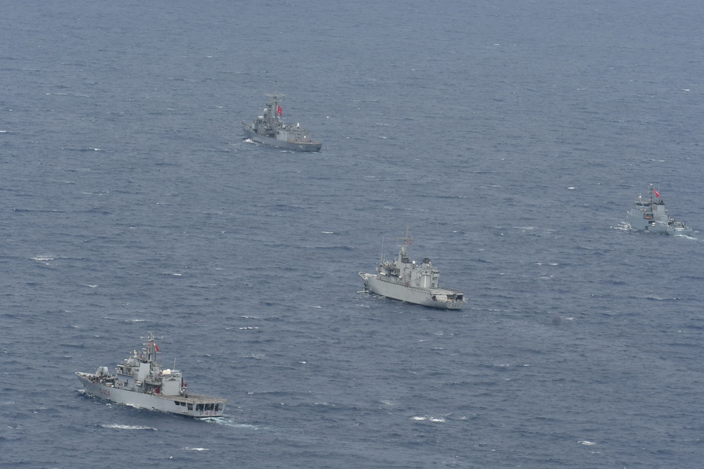 An aerial photograph shows four gray naval vessels sailing in a loose formation across open blue-gray sea. All ships appear to be military warships, each flying what appears to be a red flag, suggesting they belong to the same navy. The formation is spread across a wide area of open water with moderate wave conditions.