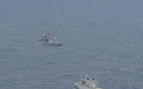 An aerial photograph shows four gray naval vessels sailing in a loose formation across open blue-gray sea. All ships appear to be military warships, each flying what appears to be a red flag, suggesting they belong to the same navy. The formation is spread across a wide area of open water with moderate wave conditions.
