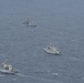 An aerial photograph shows four gray naval vessels sailing in a loose formation across open blue-gray sea. All ships appear to be military warships, each flying what appears to be a red flag, suggesting they belong to the same navy. The formation is spread across a wide area of open water with moderate wave conditions.
