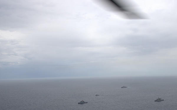 An aerial photograph taken from an aircraft showing a naval formation of multiple warships spread across open ocean waters. The ships appear to be conducting a coordinated naval exercise or transit operation under overcast skies. The image is captured through or near the aircraft's wing or fuselage edge visible at the top of the frame.