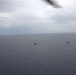 An aerial photograph taken from an aircraft showing a naval formation of multiple warships spread across open ocean waters. The ships appear to be conducting a coordinated naval exercise or transit operation under overcast skies. The image is captured through or near the aircraft's wing or fuselage edge visible at the top of the frame.