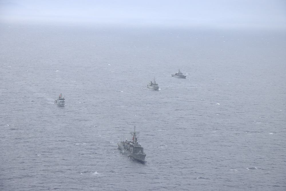 An aerial photograph shows four naval vessels sailing in a loose formation across open ocean under overcast, hazy conditions. The ships appear to be gray-painted military vessels of varying sizes, consistent with a naval exercise or patrol operation. Visibility is reduced due to fog or mist, giving the scene a muted, monochromatic appearance.
