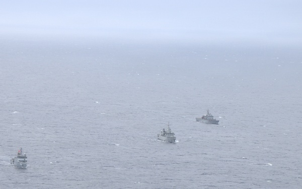 An aerial photograph shows four naval vessels sailing in a loose formation across open ocean under overcast, hazy conditions. The ships appear to be gray-painted military vessels of varying sizes, consistent with a naval exercise or patrol operation. Visibility is reduced due to fog or mist, giving the scene a muted, monochromatic appearance.