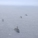 An aerial photograph shows four naval vessels sailing in a loose formation across open ocean under overcast, hazy conditions. The ships appear to be gray-painted military vessels of varying sizes, consistent with a naval exercise or patrol operation. Visibility is reduced due to fog or mist, giving the scene a muted, monochromatic appearance.