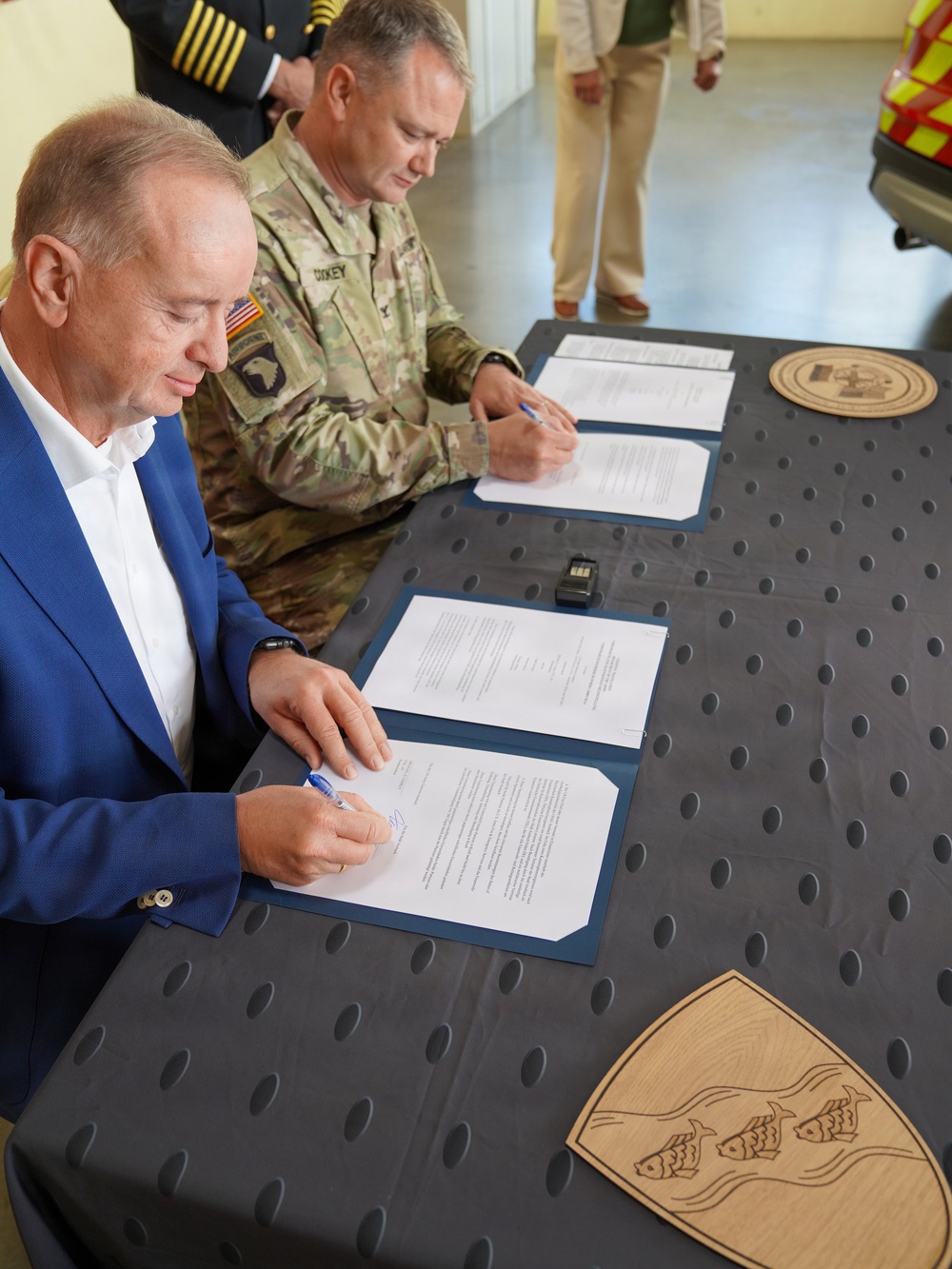 Two individuals are seated at a table signing official documents in what appears to be a formal agreement ceremony. One person is wearing a U.S. Army combat uniform with a 101st Airborne Division patch and a name tape reading 'DIKEY', while the other is in civilian attire (blue blazer, white shirt). Two wooden plaques or seals are visible on the table, and additional uniformed and civilian personnel are standing in the background.