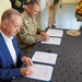 Two individuals are seated at a table signing official documents in what appears to be a formal agreement ceremony. One person is wearing a U.S. Army combat uniform with a 101st Airborne Division patch and a name tape reading 'DIKEY', while the other is in civilian attire (blue blazer, white shirt). Two wooden plaques or seals are visible on the table, and additional uniformed and civilian personnel are standing in the background.