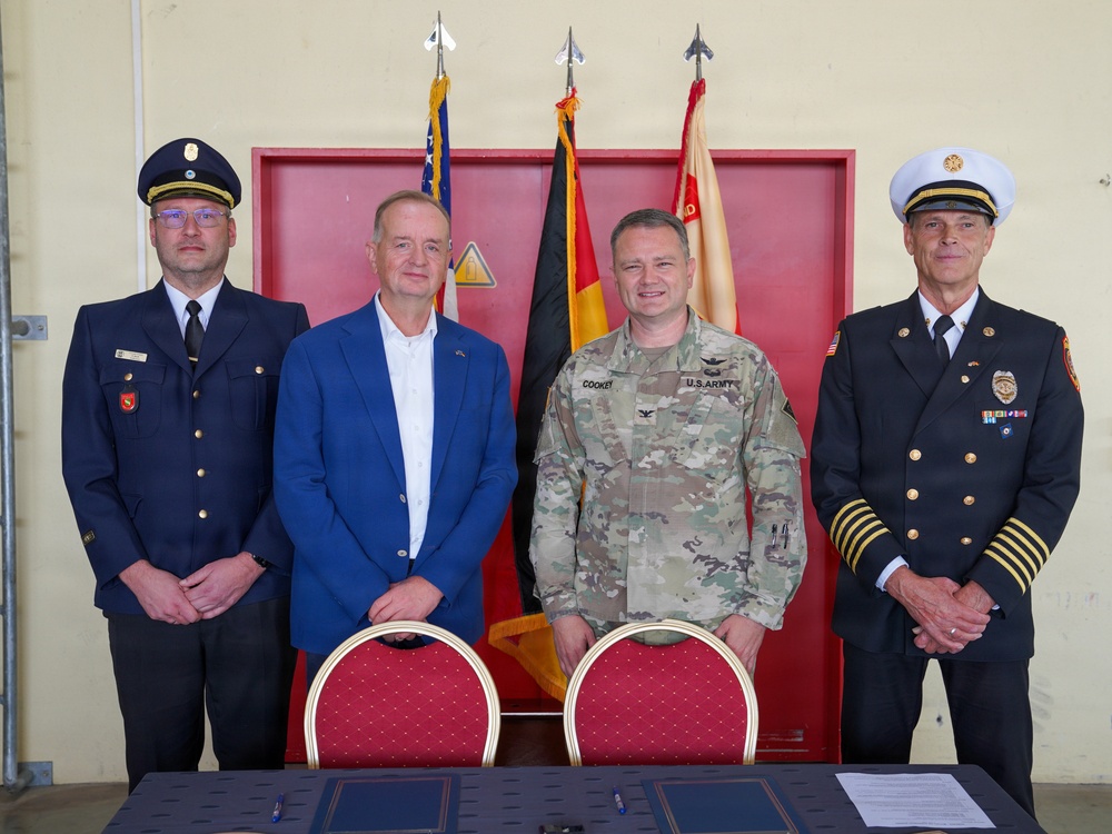 Four individuals pose for a formal group photo in what appears to be a fire station or military facility. The group includes a U.S. Army colonel (name tag reads 'COOKEY'), a civilian in a blue blazer, a German fire service officer, and a U.S. fire chief. A signing table with documents is positioned in front of them, suggesting a formal agreement or memorandum of understanding ceremony.
