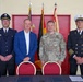 Four individuals pose for a formal group photo in what appears to be a fire station or military facility. The group includes a U.S. Army colonel (name tag reads 'COOKEY'), a civilian in a blue blazer, a German fire service officer, and a U.S. fire chief. A signing table with documents is positioned in front of them, suggesting a formal agreement or memorandum of understanding ceremony.