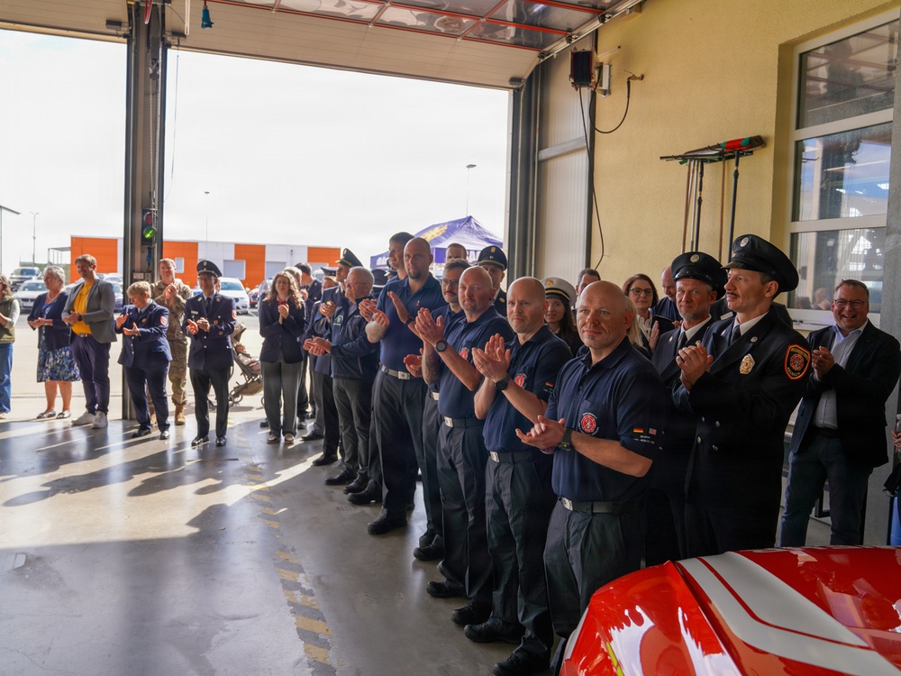 A group of firefighters and civilians are gathered inside what appears to be a fire station bay, applauding during what looks like a formal ceremony or event. The personnel are wearing navy blue uniforms with patches, while some wear formal dress uniforms with black caps. A red fire vehicle is partially visible in the foreground.