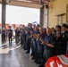 A group of firefighters and civilians are gathered inside what appears to be a fire station bay, applauding during what looks like a formal ceremony or event. The personnel are wearing navy blue uniforms with patches, while some wear formal dress uniforms with black caps. A red fire vehicle is partially visible in the foreground.