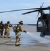 A group of firefighters in full protective gear are conducting what appears to be a training exercise involving a military helicopter, likely a UH-60 Black Hawk, on a tarmac or airfield. One firefighter is actively spraying water or foam from a hose toward the base of the helicopter, while others stand by managing the hose line. The scene suggests an aircraft rescue and firefighting (ARFF) drill.