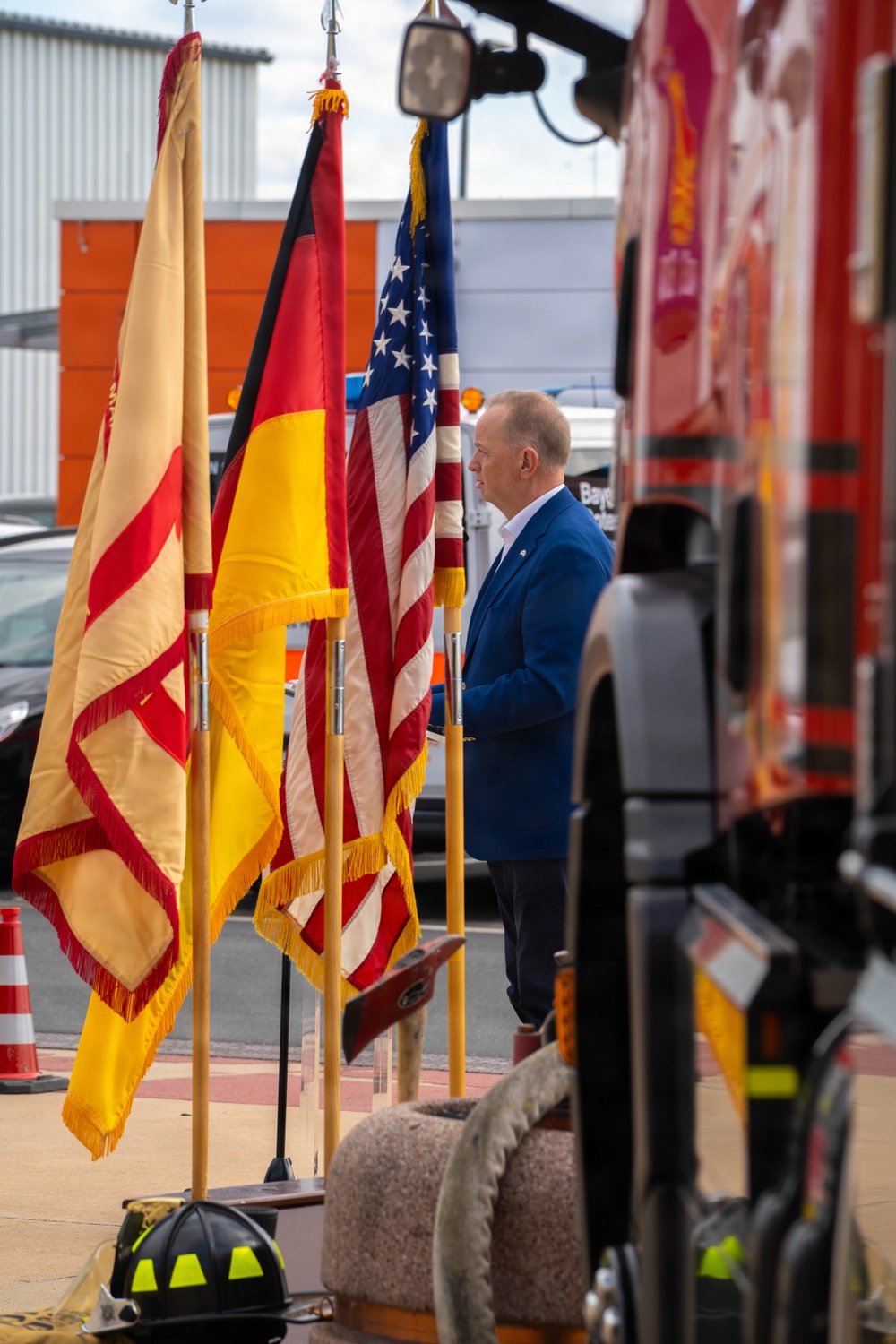 A man in a blue suit stands at attention beside a row of ceremonial flags including the German flag, the American flag, and what appears to be a regional or organizational flag with red and yellow colors. The setting appears to be an outdoor ceremony at a fire station or emergency services facility, with a red fire truck visible on the right side. A firefighter helmet is visible on the ground in the foreground.
