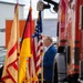 A man in a blue suit stands at attention beside a row of ceremonial flags including the German flag, the American flag, and what appears to be a regional or organizational flag with red and yellow colors. The setting appears to be an outdoor ceremony at a fire station or emergency services facility, with a red fire truck visible on the right side. A firefighter helmet is visible on the ground in the foreground.