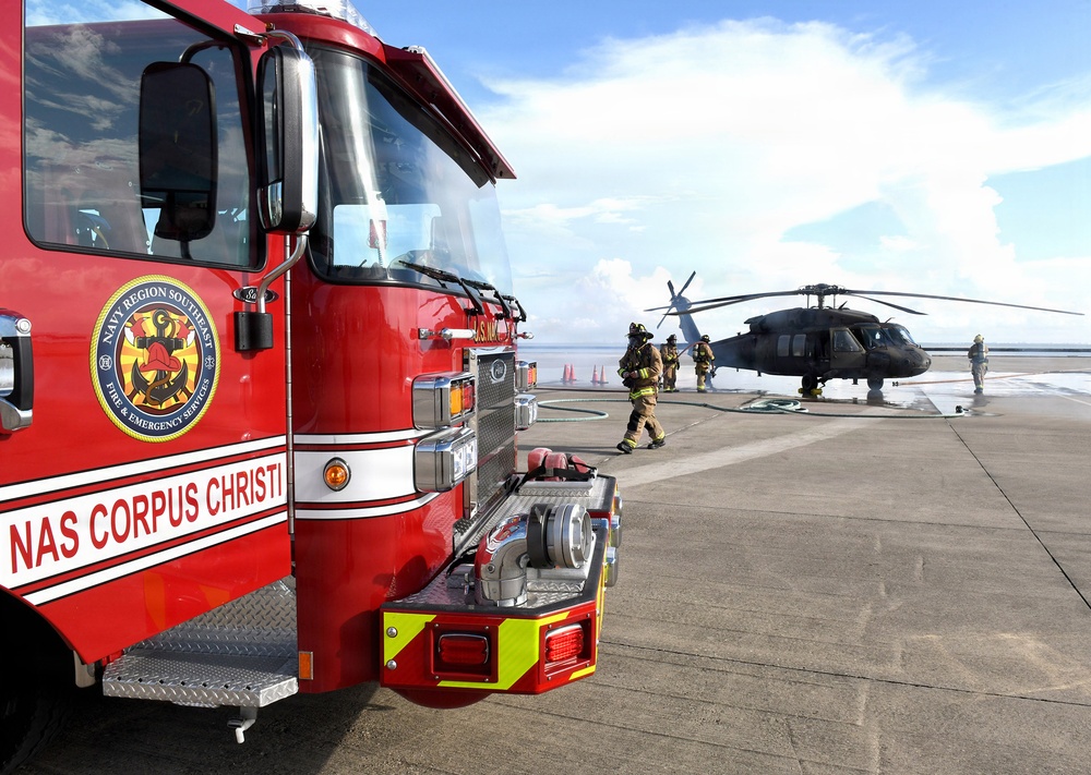 A red fire truck belonging to NAS Corpus Christi (Naval Air Station Corpus Christi) Fire &amp; Emergency Services, under Navy Region Southeast, is positioned on a tarmac or flight deck. Firefighters in full gear are responding near a military helicopter, likely a UH-60 Black Hawk, with fire hoses deployed on the ground. The scene appears to be a training exercise or emergency response drill at the naval air station.