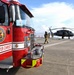 A red fire truck belonging to NAS Corpus Christi (Naval Air Station Corpus Christi) Fire &amp; Emergency Services, under Navy Region Southeast, is positioned on a tarmac or flight deck. Firefighters in full gear are responding near a military helicopter, likely a UH-60 Black Hawk, with fire hoses deployed on the ground. The scene appears to be a training exercise or emergency response drill at the naval air station.