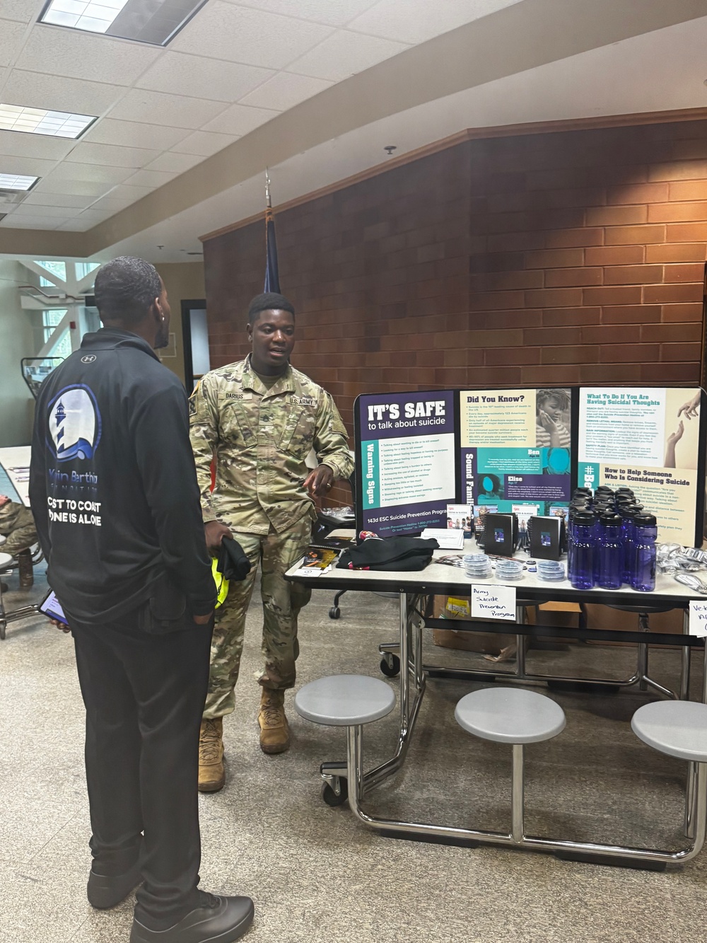 A U.S. Army soldier in camouflage uniform is staffing an informational booth for the '143d ESC Suicide Prevention Program' at what appears to be a military installation or community center. The soldier is engaged in conversation with a civilian wearing a black jacket with 'Martin Luther Foundation' branding. The display table features educational materials, blue water bottles, and signage reading 'It's Safe to talk about suicide' along with warning signs and resource information.