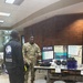 A U.S. Army soldier in camouflage uniform is staffing an informational booth for the '143d ESC Suicide Prevention Program' at what appears to be a military installation or community center. The soldier is engaged in conversation with a civilian wearing a black jacket with 'Martin Luther Foundation' branding. The display table features educational materials, blue water bottles, and signage reading 'It's Safe to talk about suicide' along with warning signs and resource information.