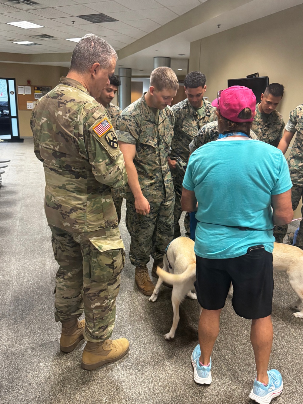 A group of military personnel in camouflage uniforms are gathered indoors around a light-colored dog, along with a civilian handler wearing a teal shirt and pink cap. The setting appears to be a military facility or administrative building with fluorescent lighting and a drop ceiling. This appears to be a therapy or comfort dog visit program for service members.