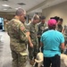 A group of military personnel in camouflage uniforms are gathered indoors around a light-colored dog, along with a civilian handler wearing a teal shirt and pink cap. The setting appears to be a military facility or administrative building with fluorescent lighting and a drop ceiling. This appears to be a therapy or comfort dog visit program for service members.