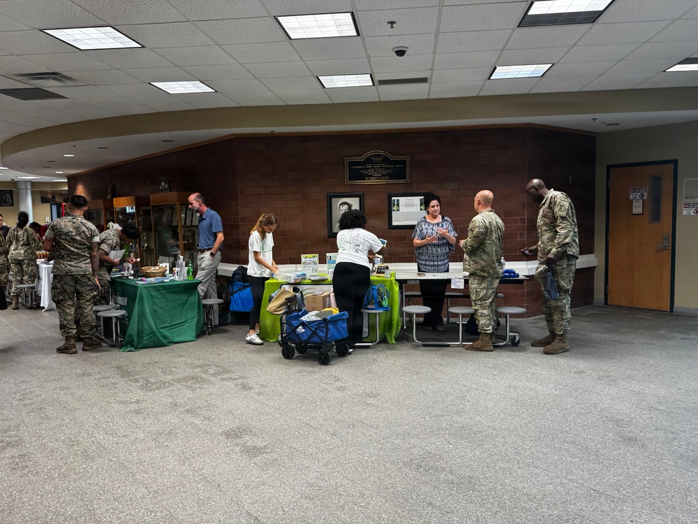 An indoor event or information fair is taking place in what appears to be a military installation building, with several vendor or resource tables set up along a brick wall. Multiple individuals in military camouflage uniforms (ACU/OCP pattern) are interacting with civilian staff at the tables. The setting resembles a community outreach, wellness fair, or resource expo hosted for military personnel.