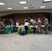 An indoor event or information fair is taking place in what appears to be a military installation building, with several vendor or resource tables set up along a brick wall. Multiple individuals in military camouflage uniforms (ACU/OCP pattern) are interacting with civilian staff at the tables. The setting resembles a community outreach, wellness fair, or resource expo hosted for military personnel.