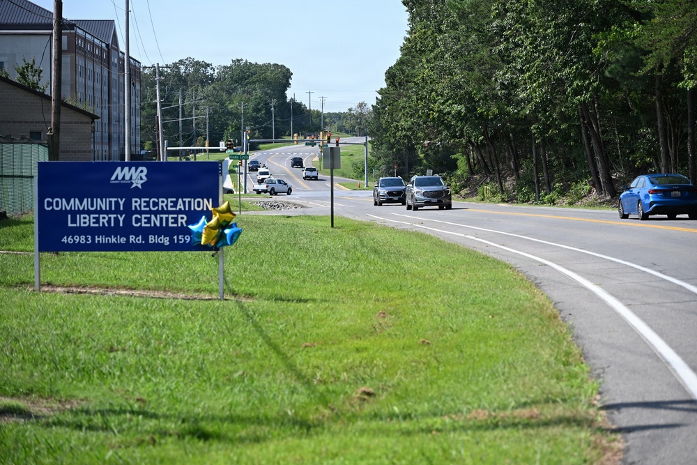 A roadside scene featuring a blue MWR (Morale, Welfare, and Recreation) sign for the Community Recreation Liberty Center located at 46983 Hinkle Rd, Building 159. The image shows a two-lane road with moderate traffic, flanked by green grass, trees, and utility poles under a clear blue sky. Decorative gold and blue star-shaped balloons are attached to the sign, suggesting a recent opening or special event.