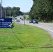 A roadside scene featuring a blue MWR (Morale, Welfare, and Recreation) sign for the Community Recreation Liberty Center located at 46983 Hinkle Rd, Building 159. The image shows a two-lane road with moderate traffic, flanked by green grass, trees, and utility poles under a clear blue sky. Decorative gold and blue star-shaped balloons are attached to the sign, suggesting a recent opening or special event.